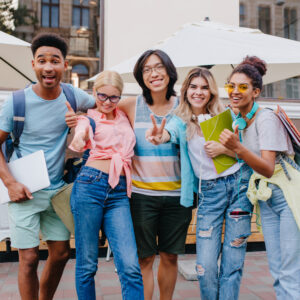 Happy blonde girl wears jeans with holes posing outdoor near smiling friends. Outdoor portrait of pleased students holding laptop and backpacks in morning.